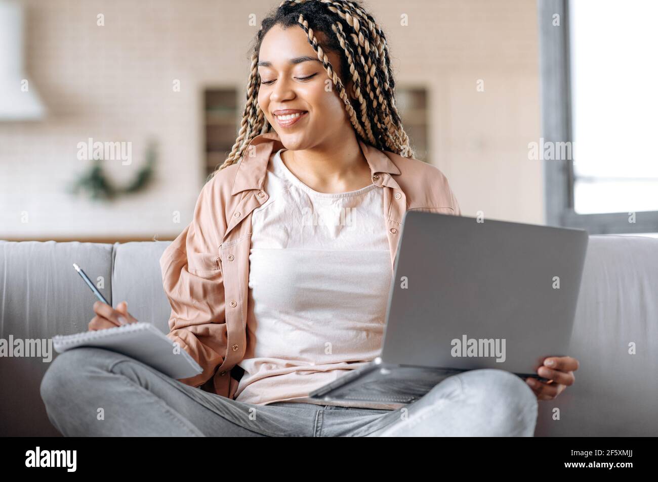 Joyful mixed race black young woman with dreadlocks, in casual wear, learns remotely from home, sits on the couch, uses a laptop, takes notes in the notebook, smiling gladly Stock Photo