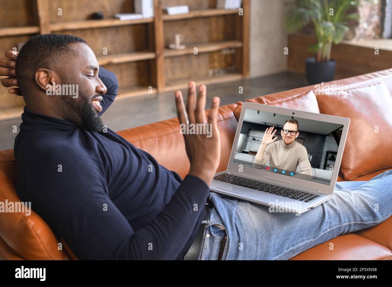 Photo over shoulder of black unshaved man having video call, greeting, waving hand, using laptop app, fast and easy communication concept, talking with best male friend, friendship through the ages Stock Photo