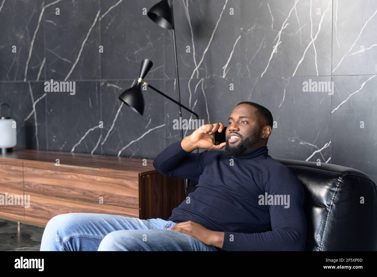 Portrait of handsome mature unshaved man with African ancestry sitting in modern office, dressed in casual comfort clothes, talking on mobile phone with business partner, communication concept Stock Photo