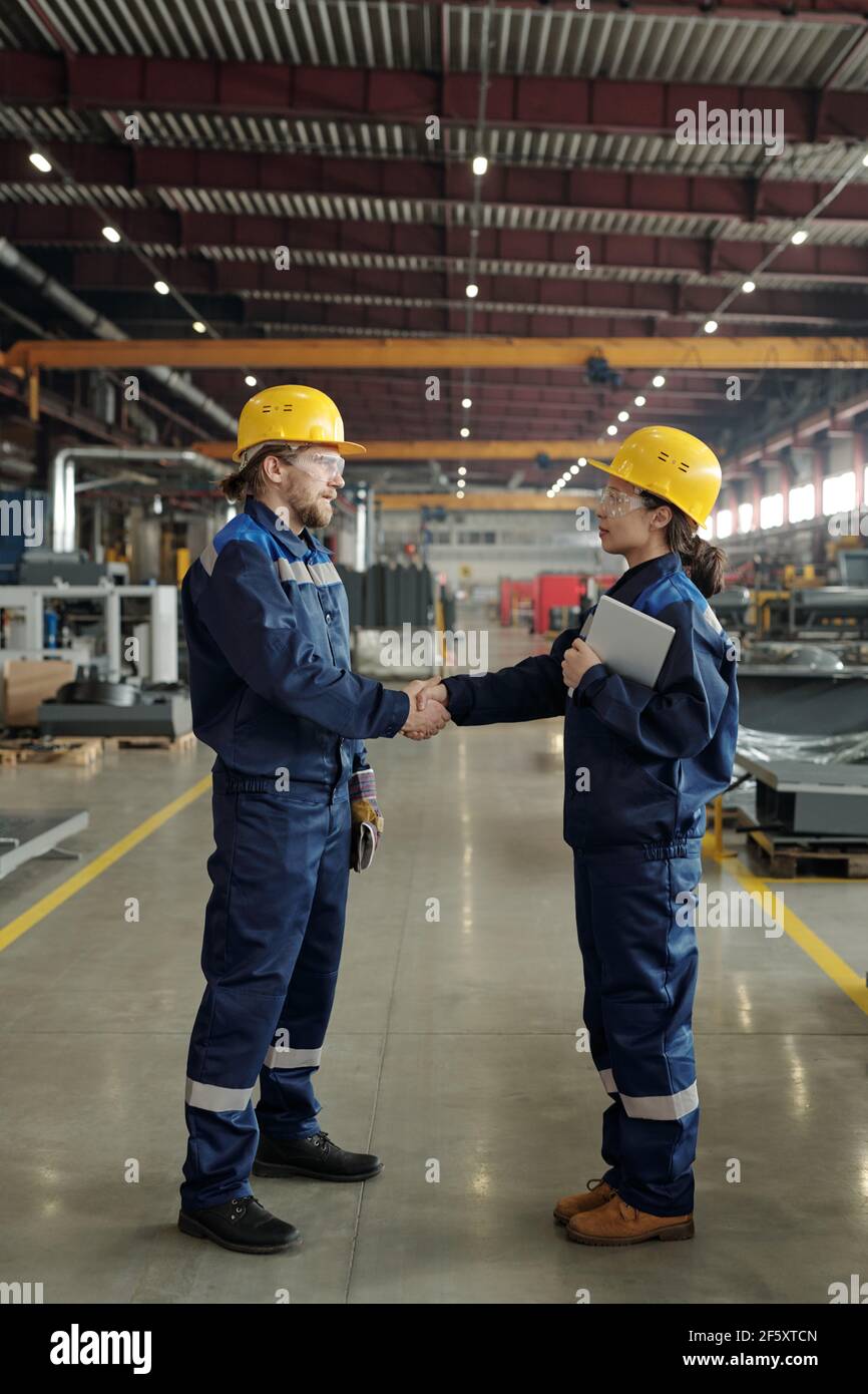 Two young successful engineers in protective hardhats and workwear looking at one another while handshaking inside large workshop Stock Photo