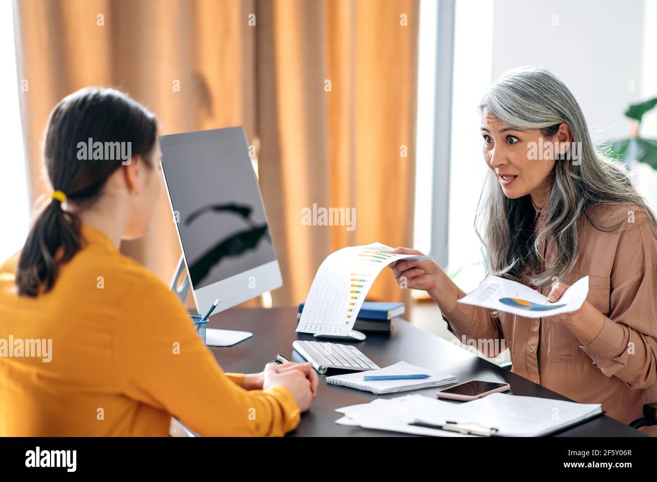 Angry indignant mature Asian woman, business leader, boss, top manager, in her office scolds an employee for a poorly drawn up business report, gestures with her hands, holds documents Stock Photo
