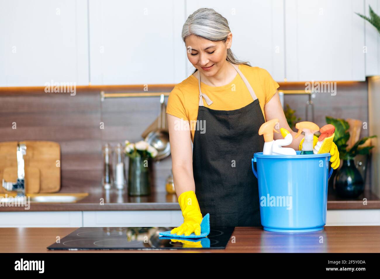 Beautiful senior gray-haired Asian cleaning lady or housewife in an apron  and gloves stands in the kitchen, wiping the stove, using a bucket of  detergents, smiling. Cleaning services Stock Photo - Alamy