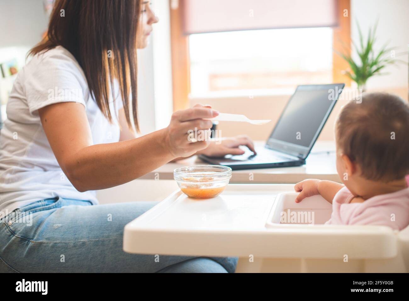Mother feed her little daughter with a spoon and search on internet food recipes Stock Photo