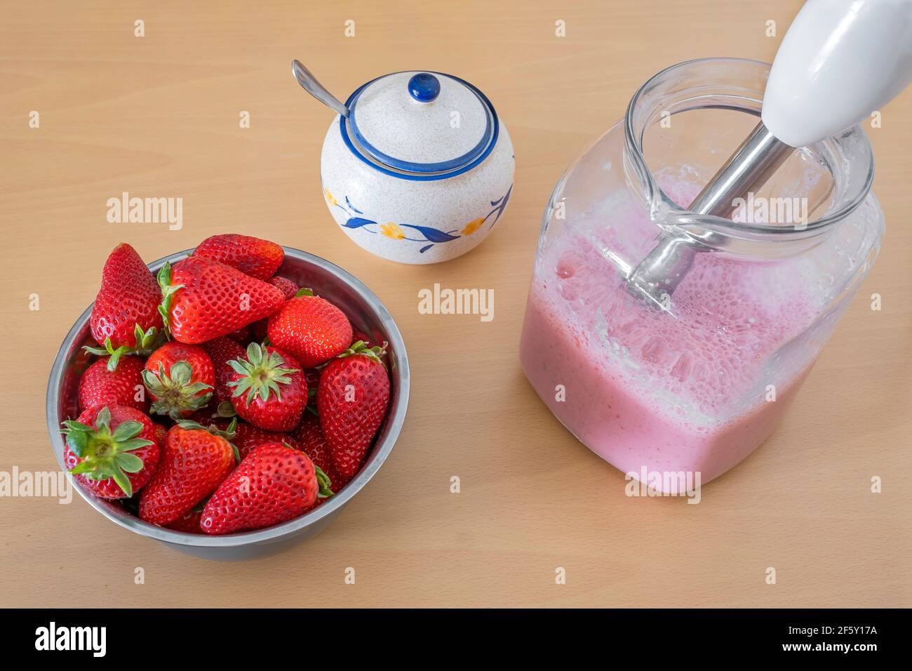 bowl full of strawberries and a pitcher with freshly made strawberry milkshake, a sugar bowl with a spoon inside, a hand blender in the pitcher Stock Photo