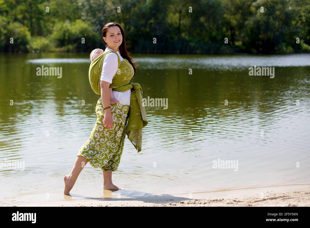 Mother with wrapping infant baby in green sling back at nature Stock Photo