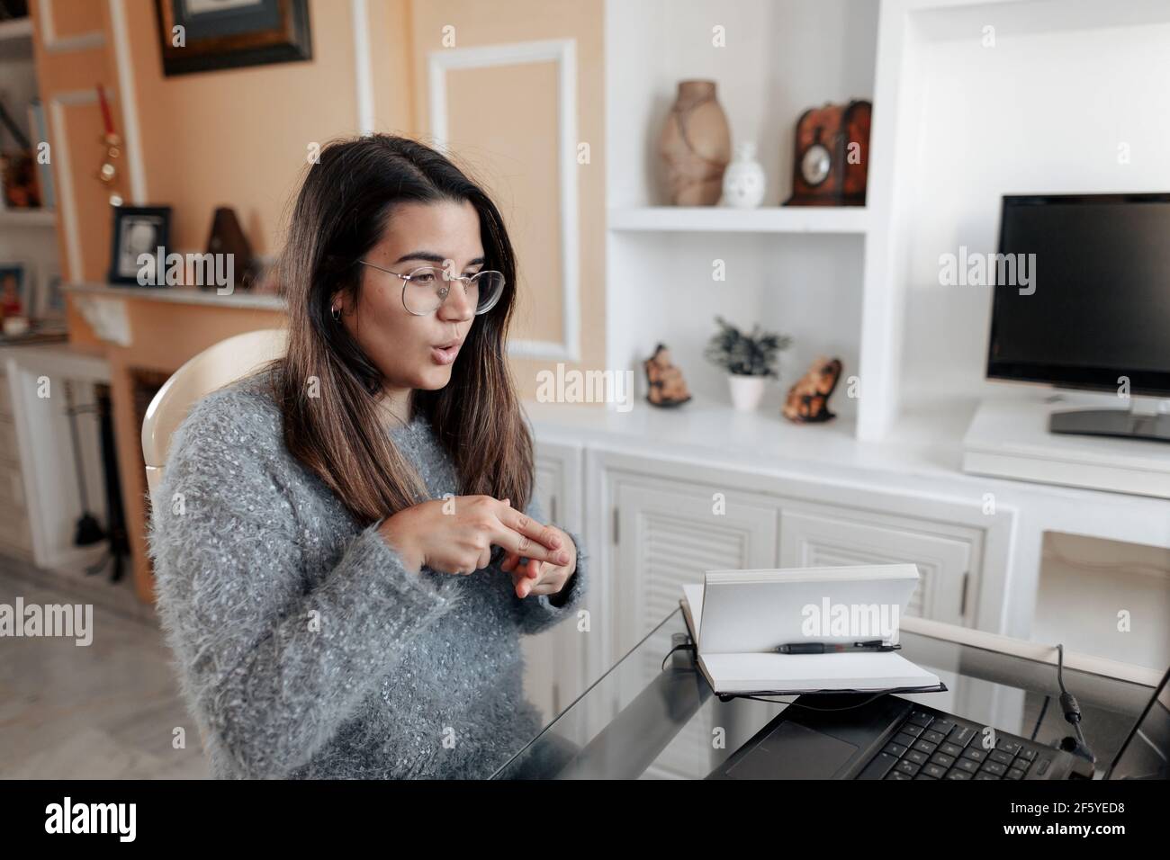 Medium shot young deaf woman wearing glasses and a sweater is using sign language to spell name while in an online video call in her living room. Stock Photo