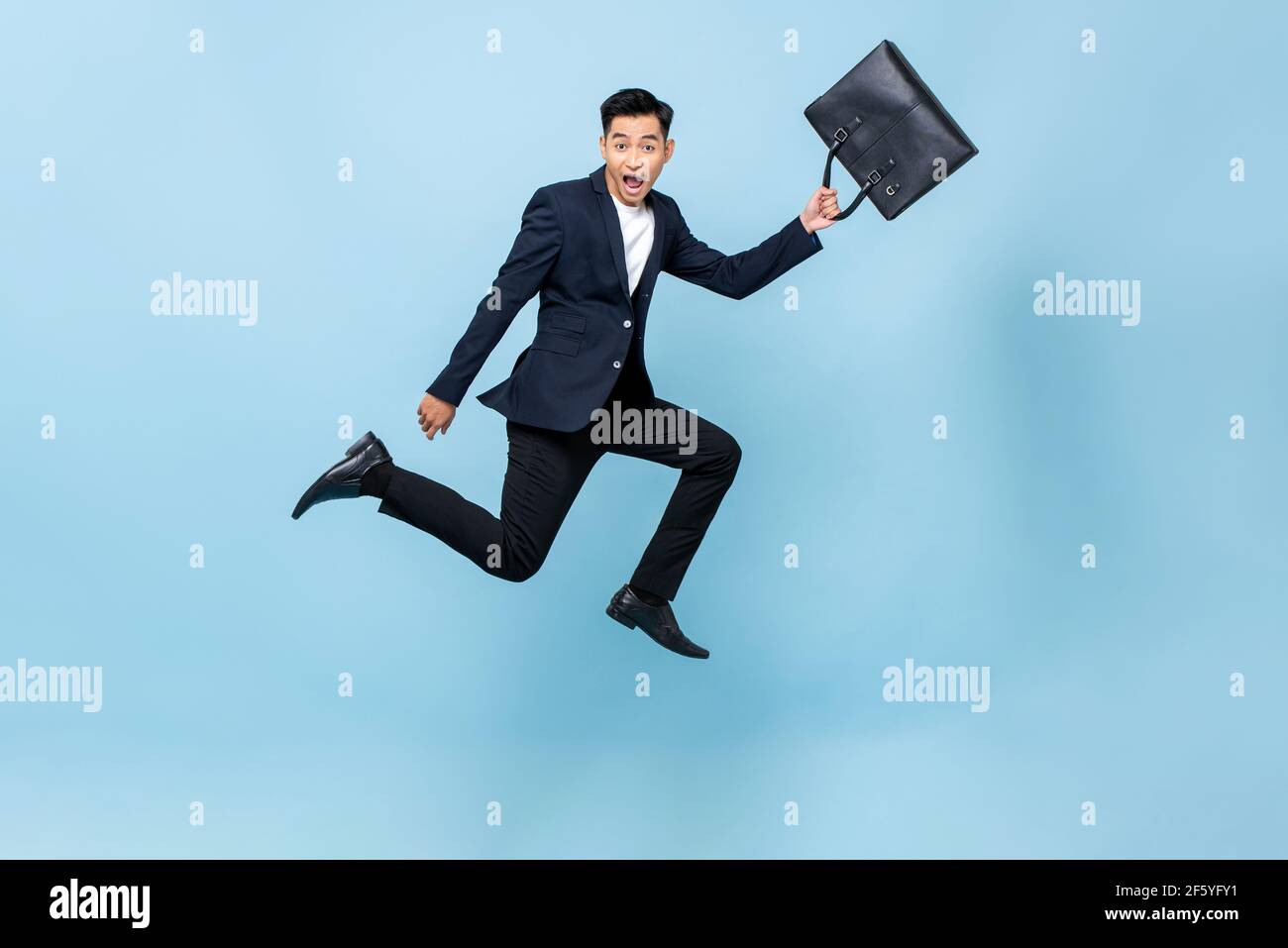 Surprised young Asian man in business suit holding bag and jumping in light blue isolated studio backkground Stock Photo