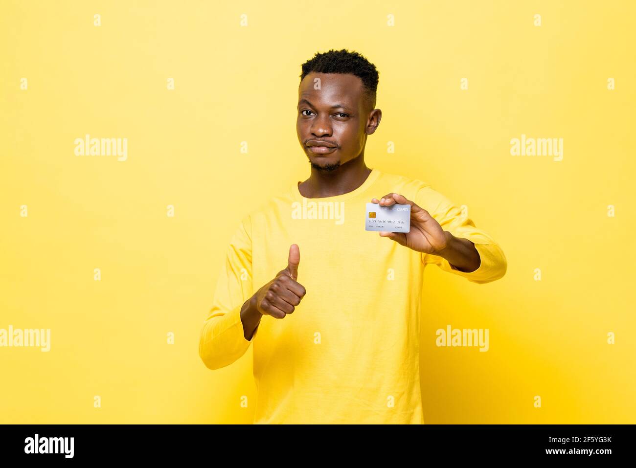 Confident African man in casual attire holding credit card and giving thumbs up on isolated yellow studio background Stock Photo