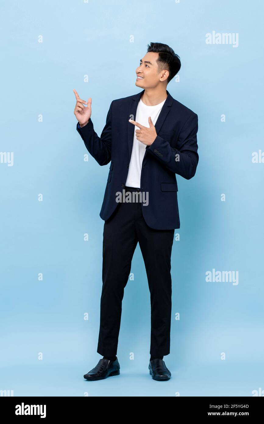 Full length portrait of happy smiling young handsome southeast Asian businessman pointing and looking upward on light blue studio background Stock Photo