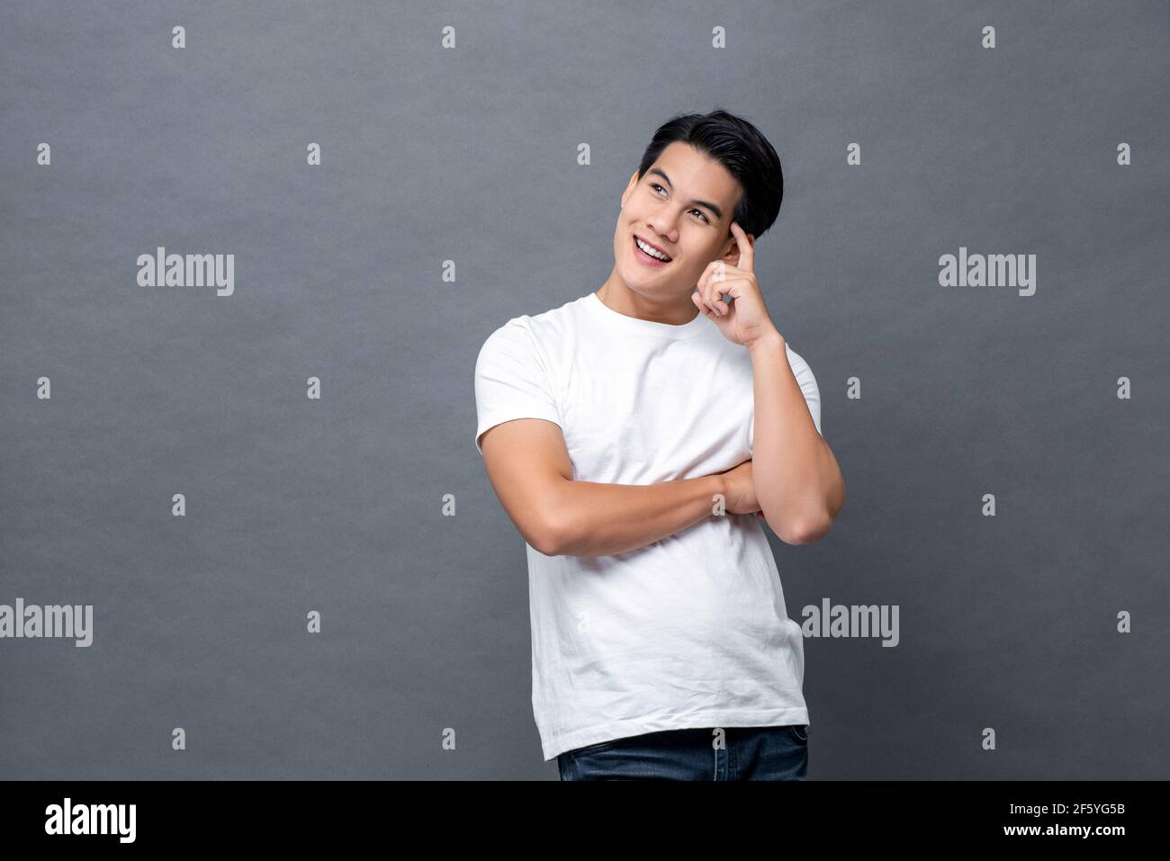 Handsome smiling friendly Asian man in casual white plain t-shirt thinking with finger on head and looking upward to copy space on gray isolated backg Stock Photo