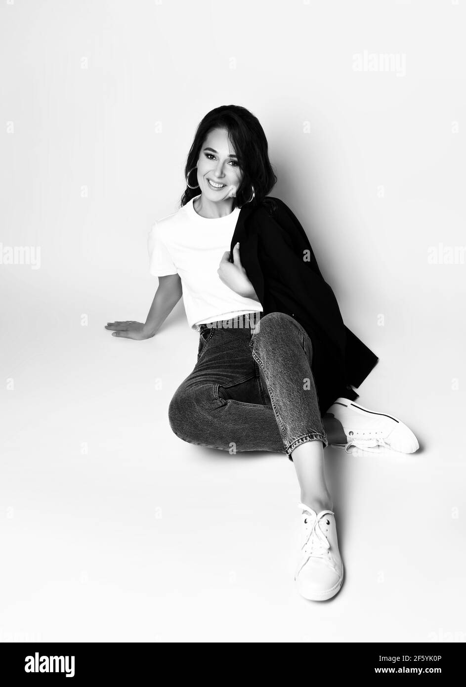 Black and white portrait of positive adult brunette woman in casual clothing t-shirt, jeans and jacket sitting on floor Stock Photo