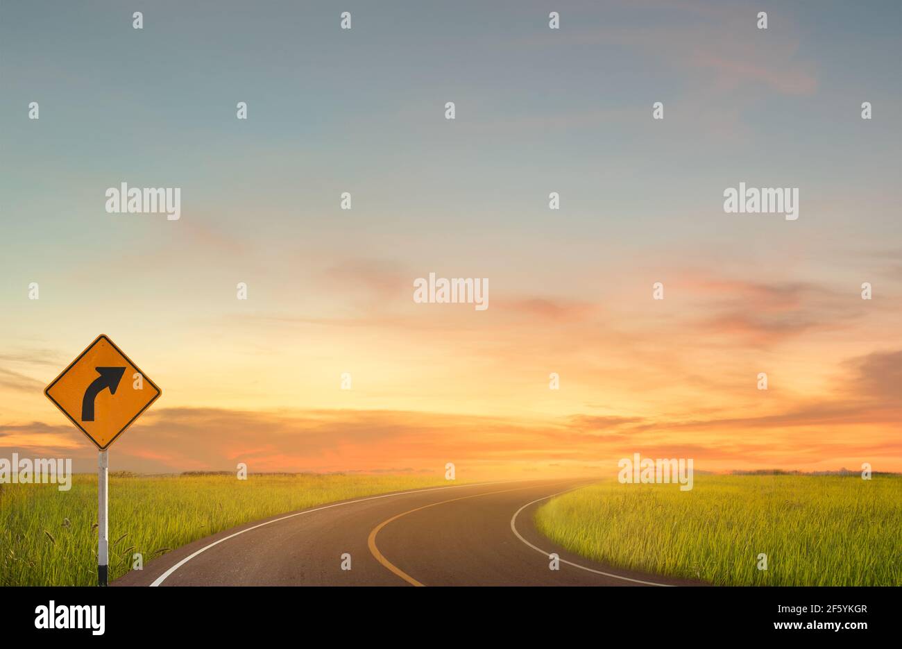 Sign curved road on the way at the natural sunset over field or meadow. Countryside Landscape Under Scenic Colorful Sky At Sunset. Nature Concept. Stock Photo