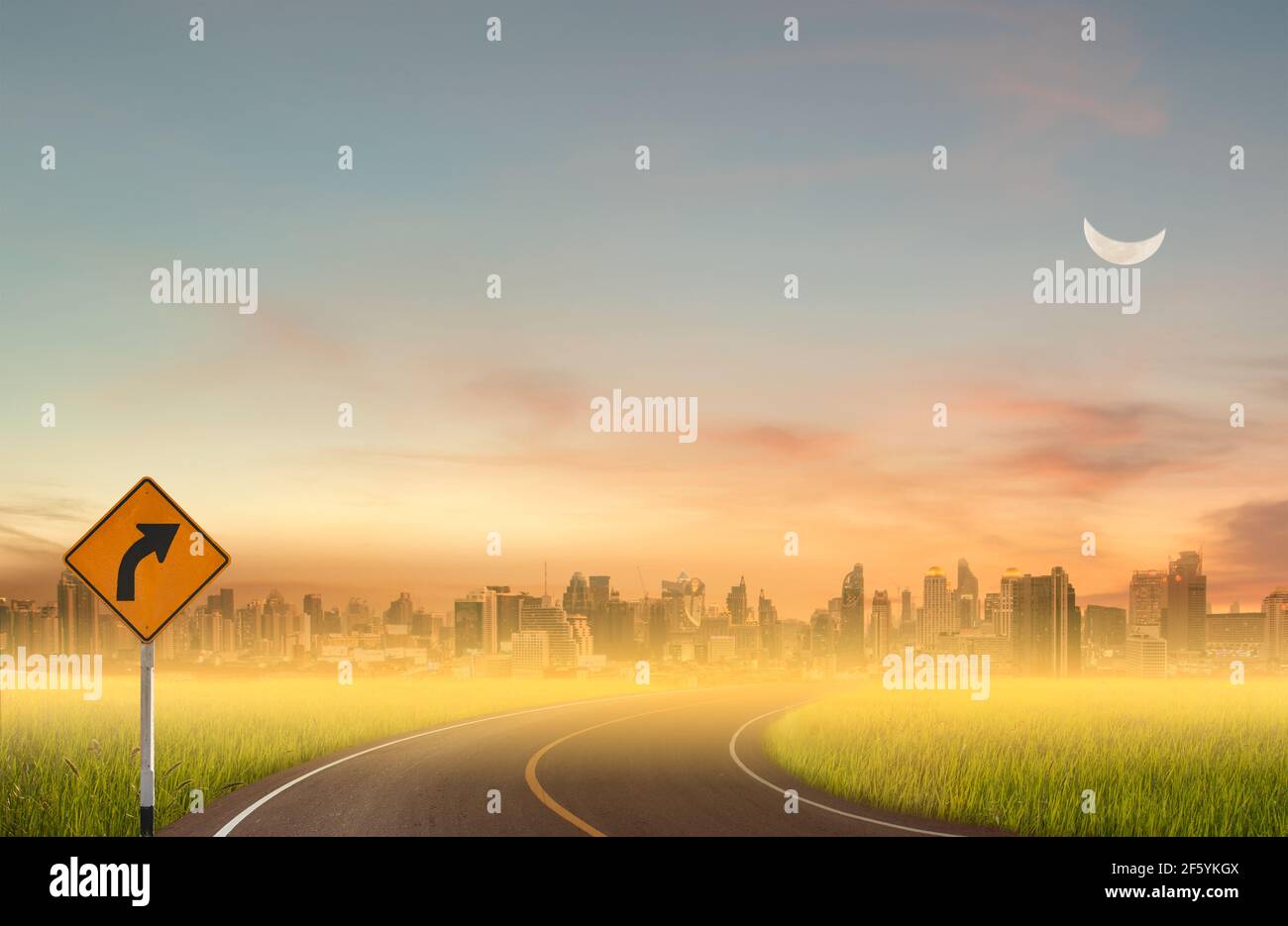 Sign curved road on the way at the natural sunset over field or meadow with moon and city background. Stock Photo