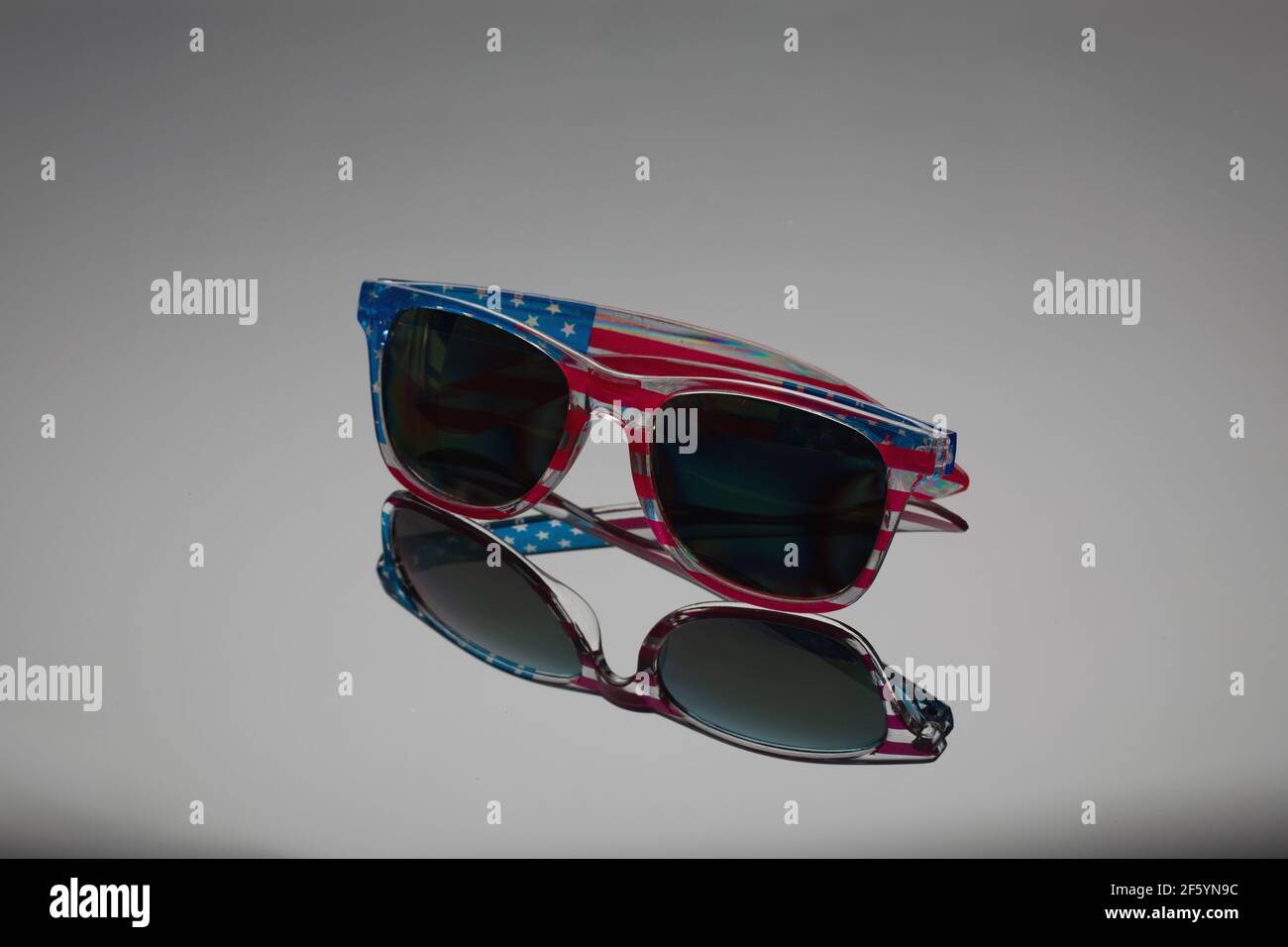 Sun safety glasses with dark glasses in frame with print of flag of America are reflected on gray mirror surface of background. Travel to United State Stock Photo