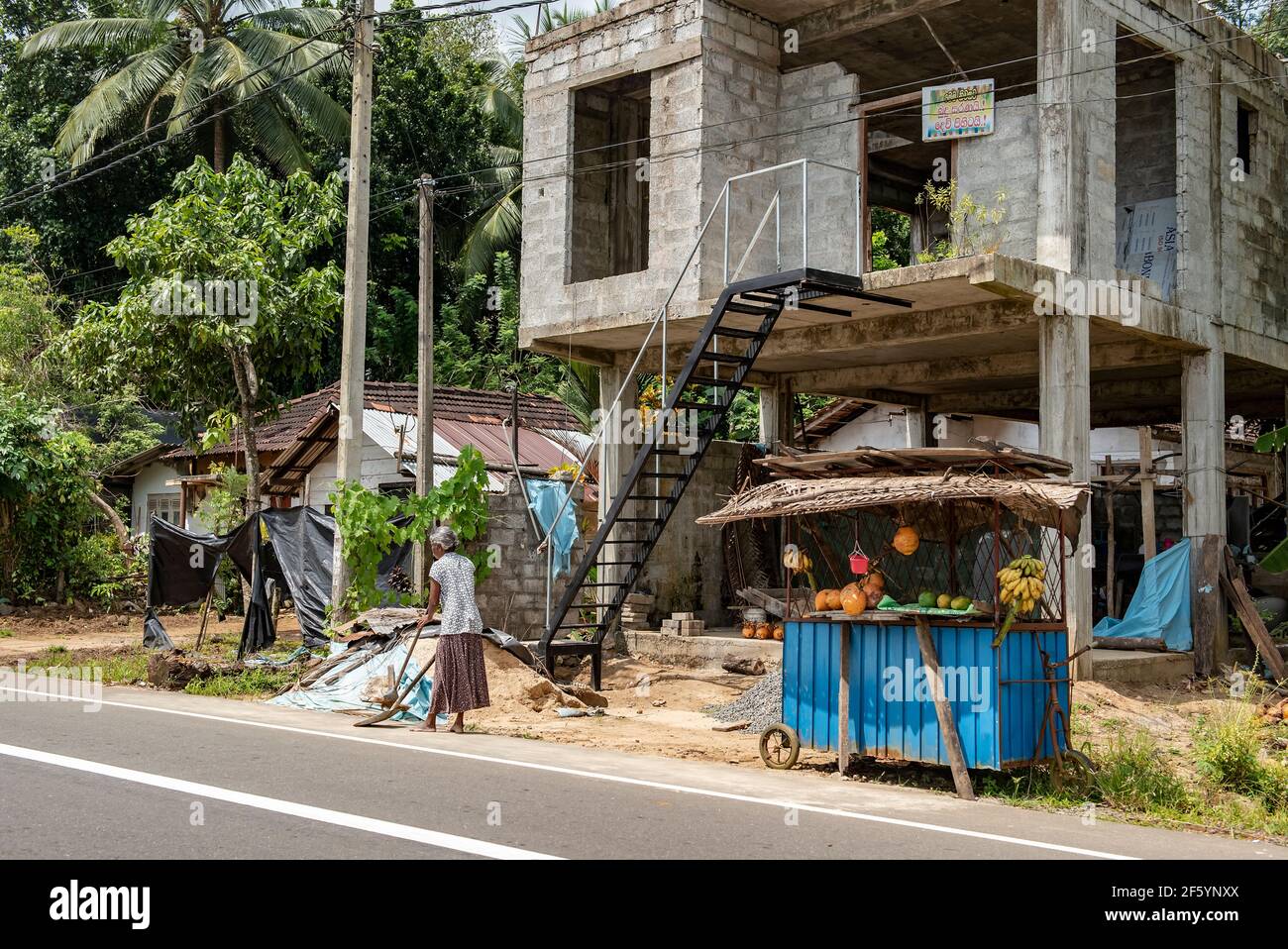 BENTOTA, SRI LANKA - 13 NOVEMBER, 2019: A woman sweeping a street and a fruit shop near an unfinished house on the island of Sri Lanka Stock Photo