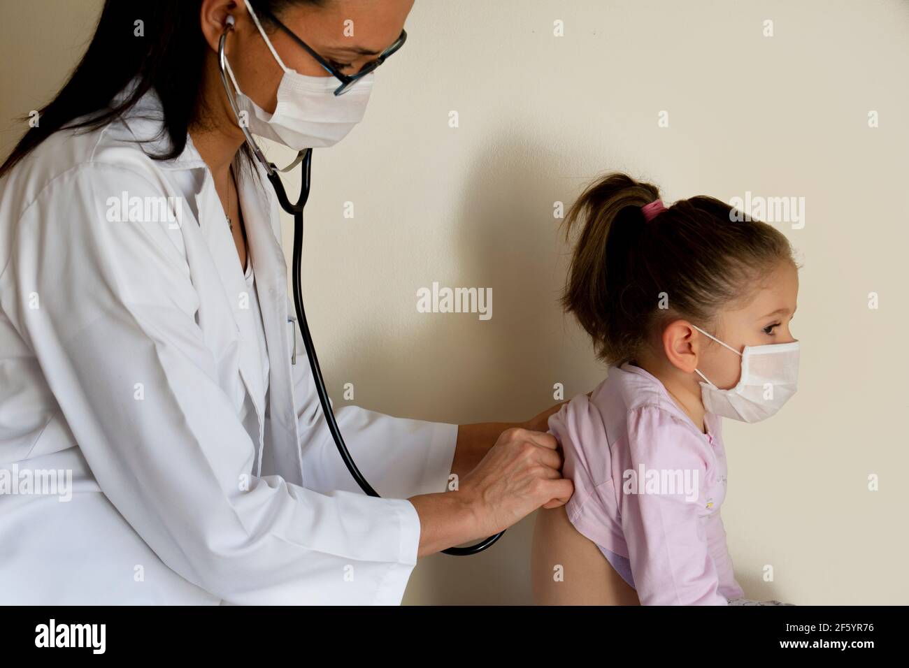 chubby little girl in pediatric examination by her doctor. the doctor listens to your lungs and heart. they wear protective face masks Stock Photo