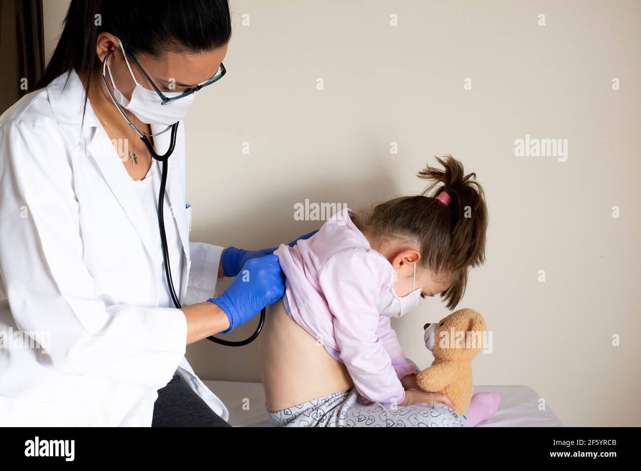 chubby little girl in pediatric examination by her doctor. the doctor listens to your lungs and heart. they wear protective face masks Stock Photo