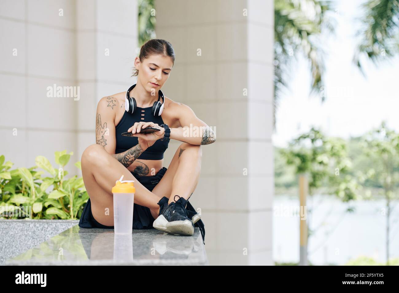 Beautiful fit young woman sitting outdoors, resting after training and checking smartwatch to control her heart rate Stock Photo
