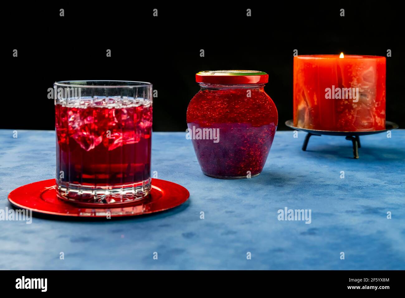 A calming glass jar filled with red glitter between a glass of red soda and a lit red candle Stock Photo