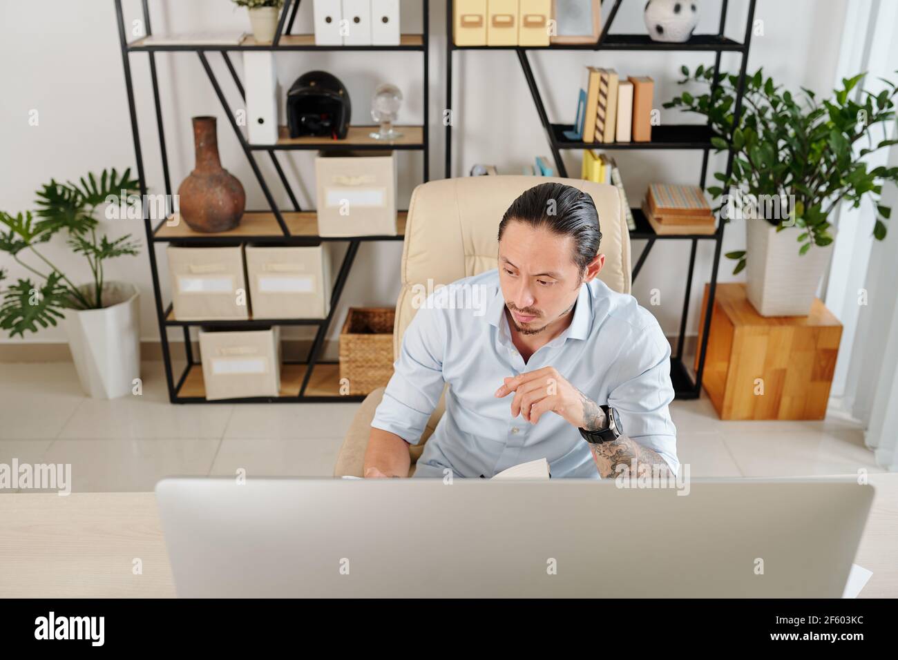 Serios frowning mixed-race entrepreneur working at desk in his home office and checking planner Stock Photo