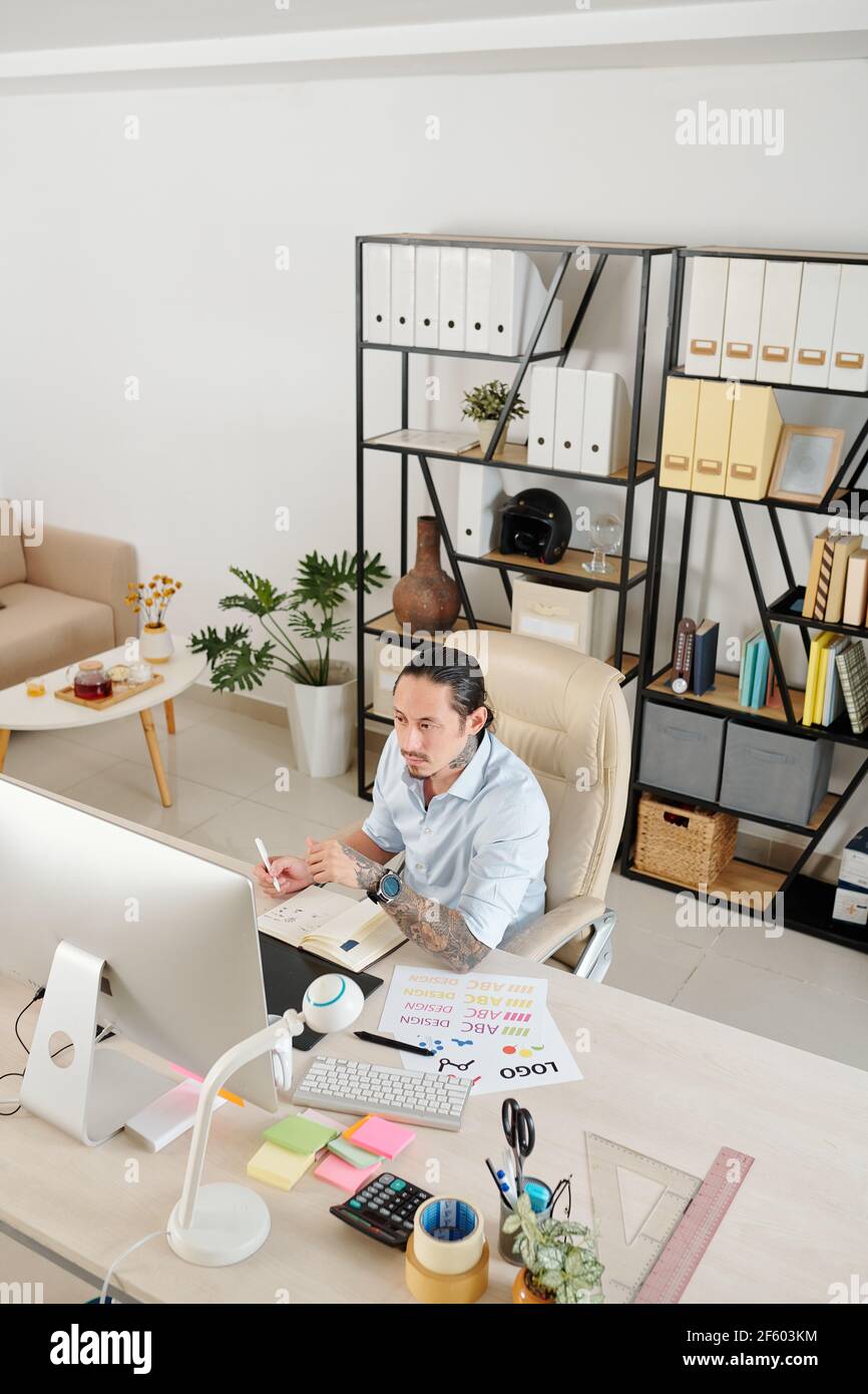 Pensive serious graphic designer working at home office, checking e-mail on computer screen and taking notes in planner Stock Photo