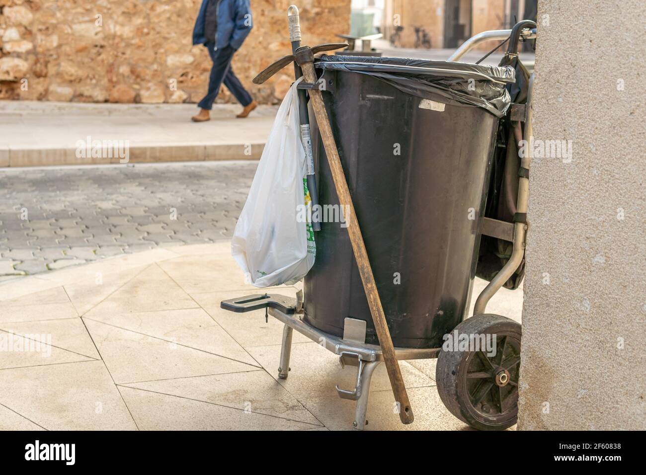 Street sweeper's cart with bucket and broom on an urban street at dawn. In the background unrecognizable pedestrians Stock Photo