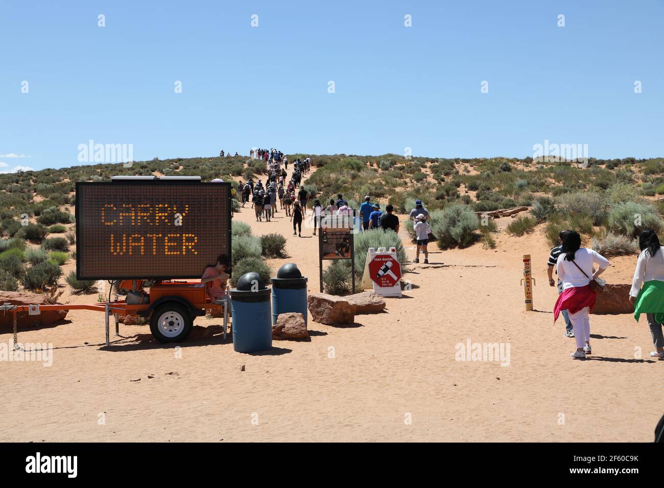 Procession of people passing a sign that warns to carry water; they are nevertheless following each other towards the horizon; global warming concept Stock Photo