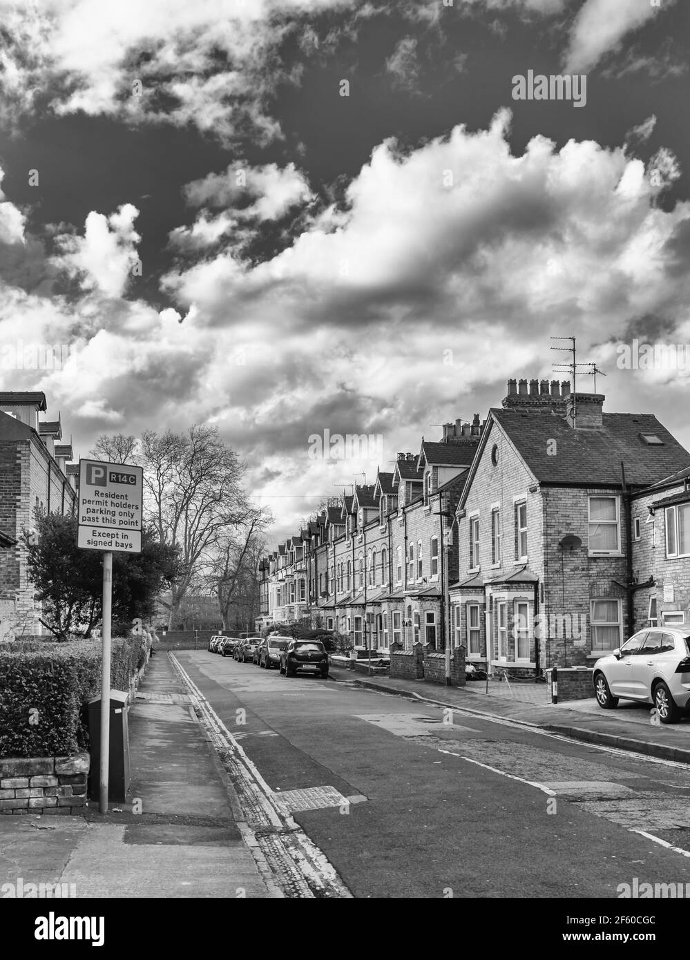 A typical city street with terraced houses made of brick and with tiled roofs.  There is a parking sign in he foreground and cars line the street. Stock Photo