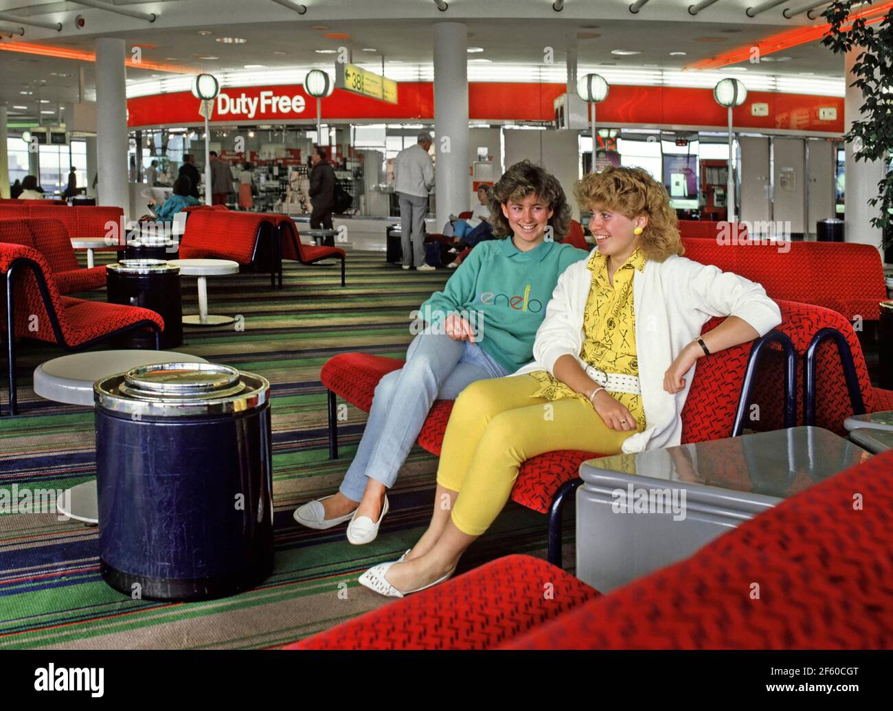 Two model released teenage girls talking and wearing 1980s fashion about to travel to a 80s summer holiday in Spain waiting inside the departure lounge building with Duty Free shop & sign beyond at Gatwick Airport West Sussex England UK an historical archive image of the way we were in 1986 Stock Photo