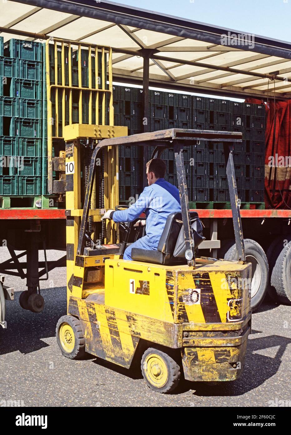 Close up 1987 historical archive image of 80s forklift truck & driver worker working on loading pallet of bottles of soft drinks in plastic crates onto articulated lorry trailer transport with open side sliding easy access curtain parked outside1980s manufacturing factory business in an archival view of the way we were in 80s Essex England UK Stock Photo