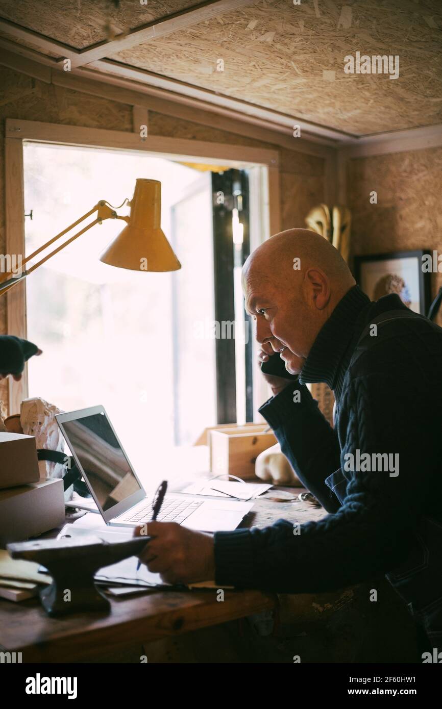 Businessman talking over mobile phone while using laptop in workshop Stock Photo