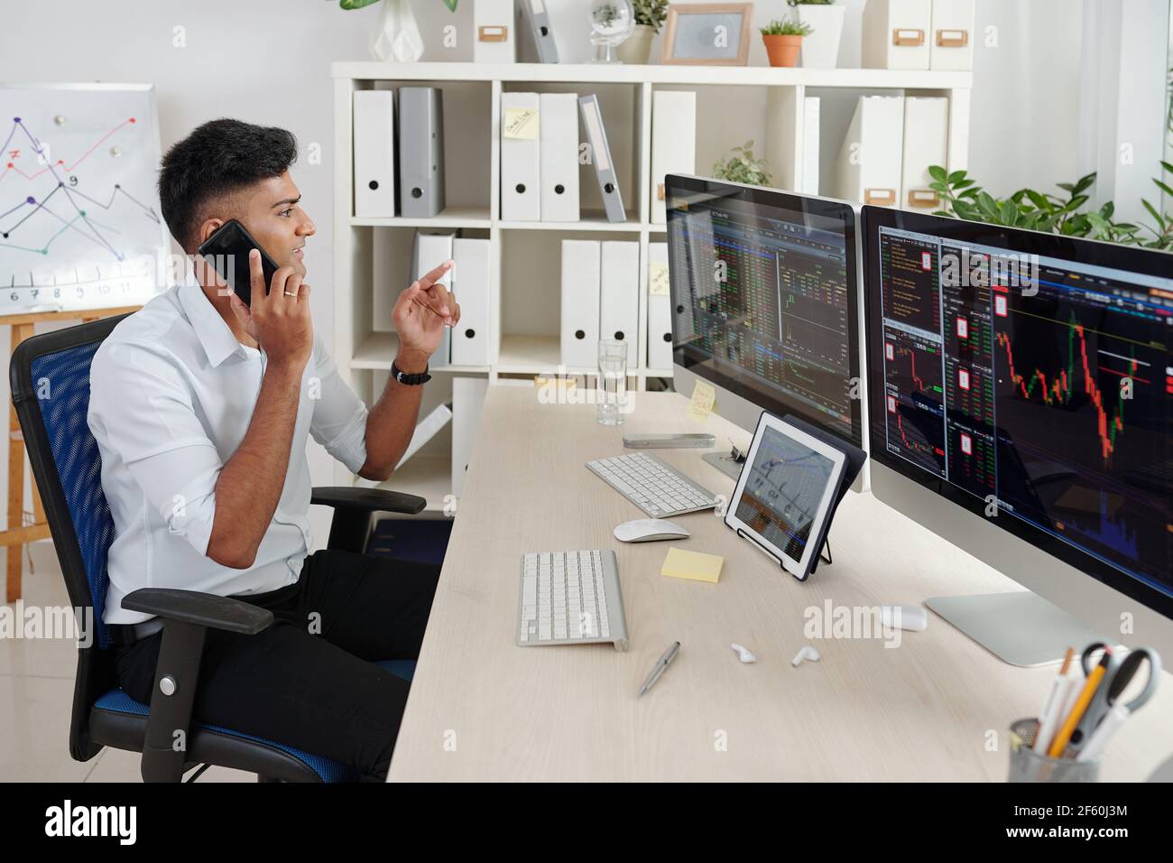Positive Indian trader analyzing stock data on computer screens and consulting clients on phone Stock Photo