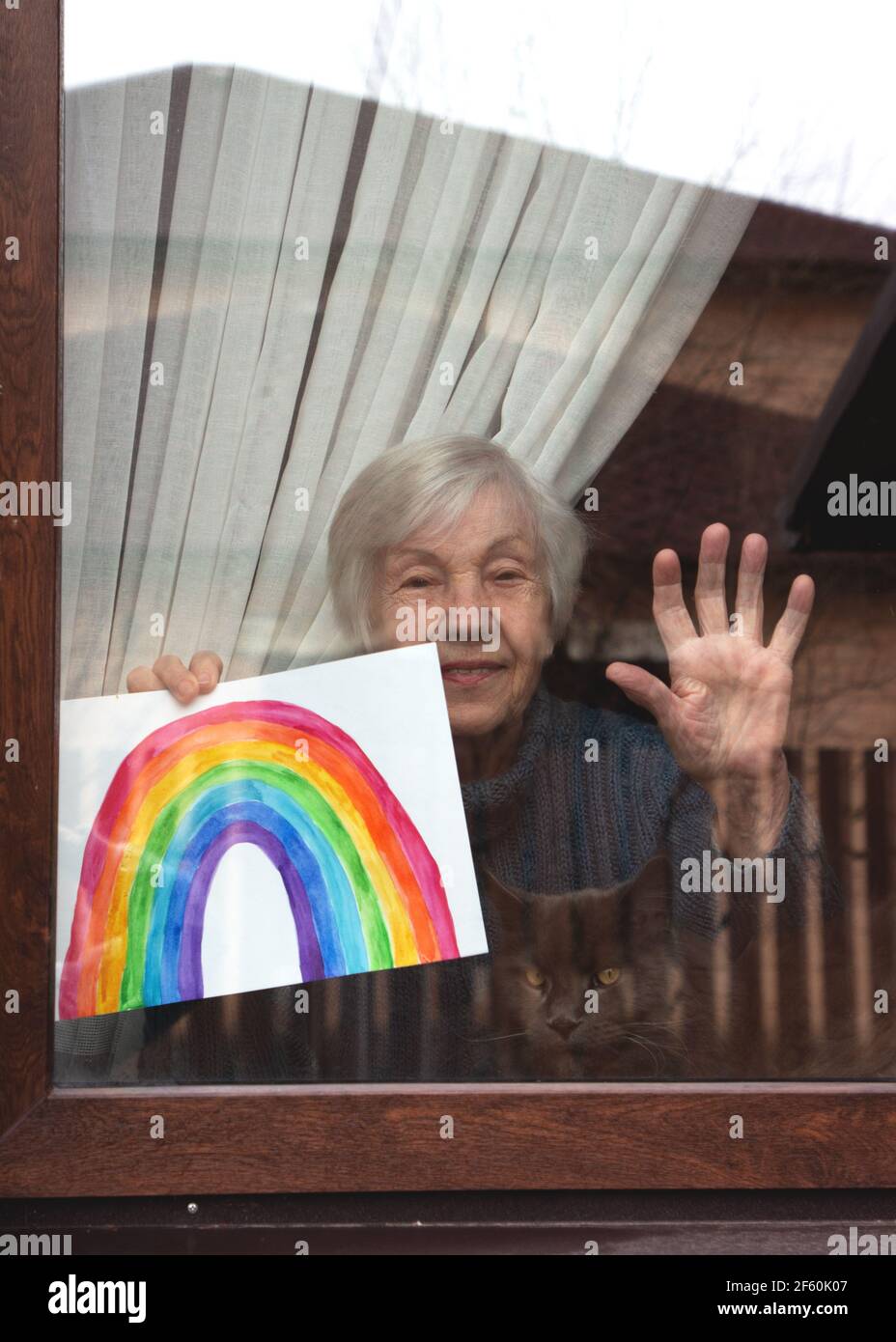 Senior woman in the window of the house holds a child's drawing of a rainbow in her hands. Rainbow in the window.Thank you NHS, Promotion in support Stock Photo