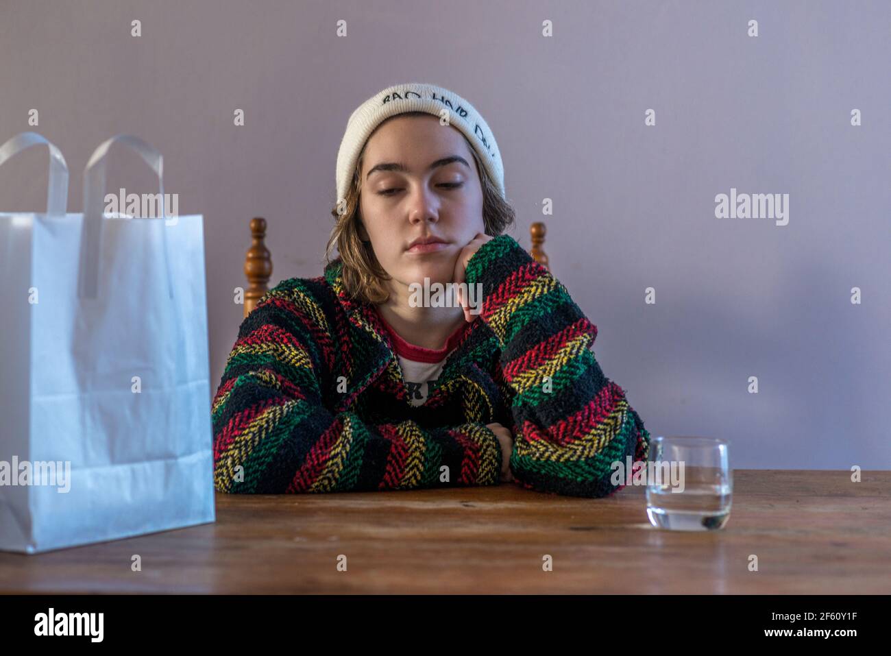 Grumpy teenage girl sitting at table in a white beanie with a glass of ...