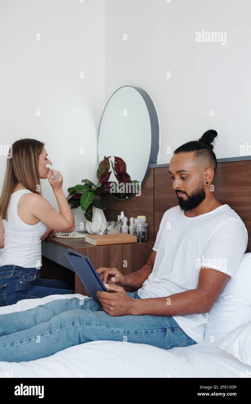Serious man sitting on bed and reading article on tablet computer when his girlfriend doing make-up at her vanity Stock Photo