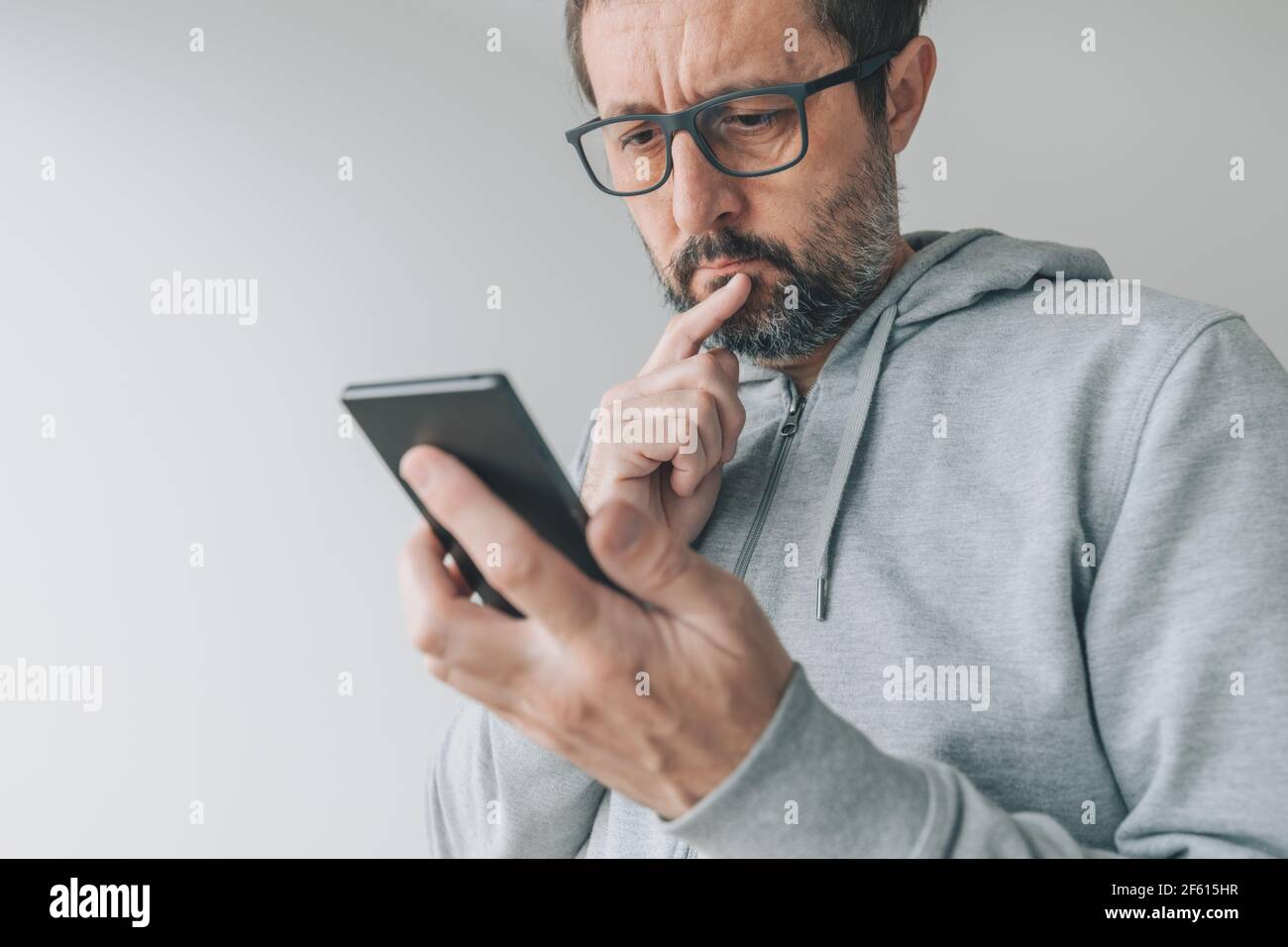 Serious thoughtful mid-adult male looking at mobile smart phone screen with finger on lips and reading text message, close up with selective focus Stock Photo