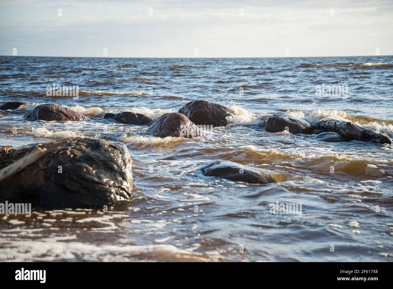 Rocks on the sea shore washed by the blue water of the sea and hit the rocks creating white splashes of water foam. Spring landscape on a warm day wit Stock Photo