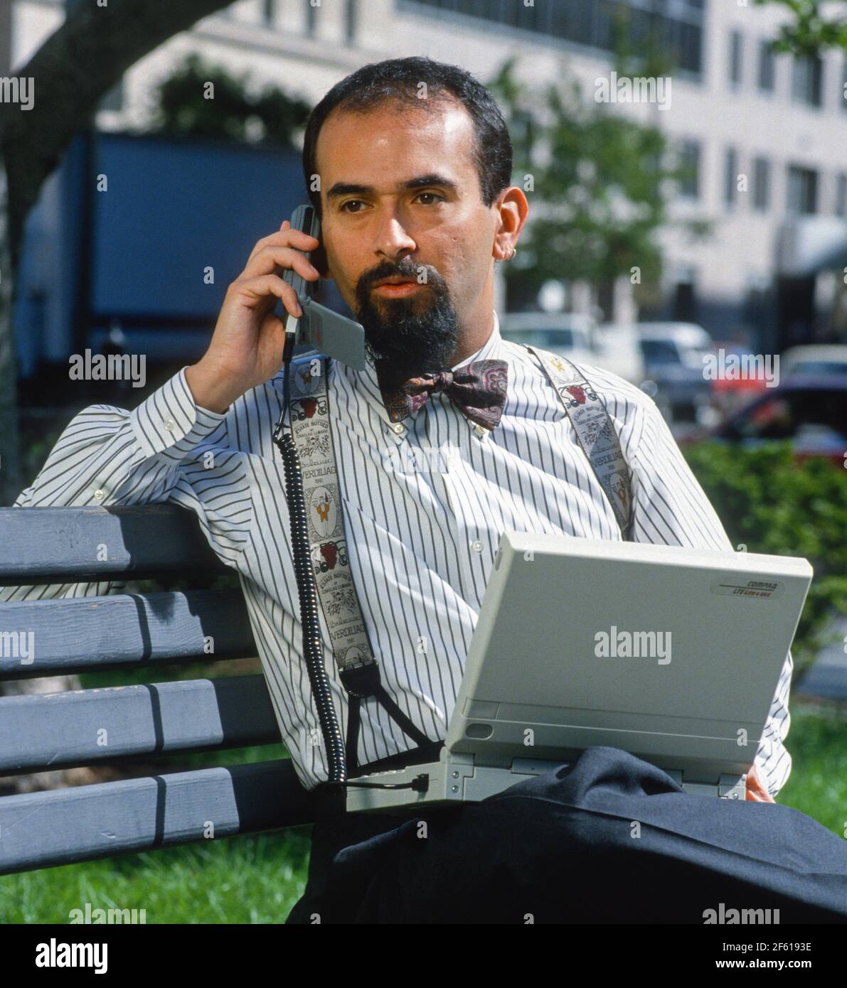 WASHINGTON, DC, USA - Man talks on mobiloe phone attached to Compaq computer using wireless modem, on park bench. Stock Photo