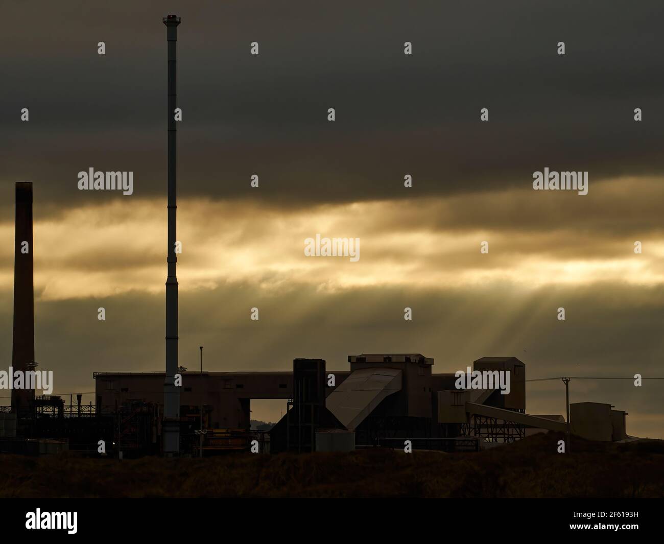 Dramatically lit, low key image of a Teeside industrial facility in near-silhouette, with a threateningly-clouded sky, broken by “god-light” sun beams Stock Photo