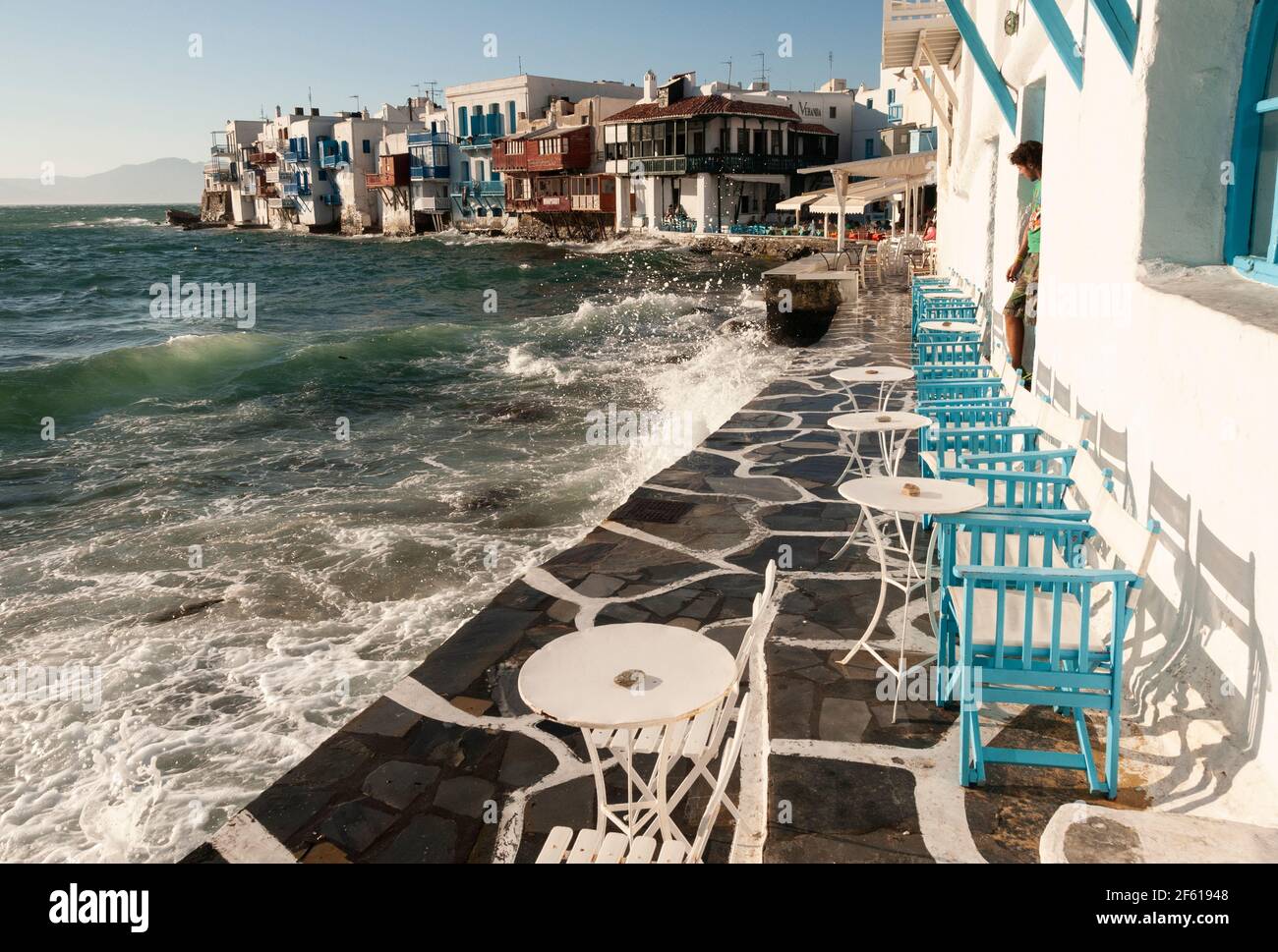 Rough sea waves splash the sea terrace of a bar in Mykonos village Greece. A man looks out of a door at the sea. Holiday travel concept Stock Photo