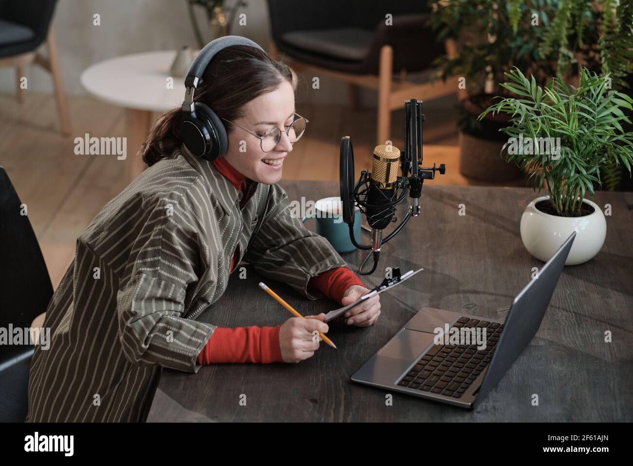 Female journalist sitting at the table in front of laptop and doing online interview on it in the studio Stock Photo