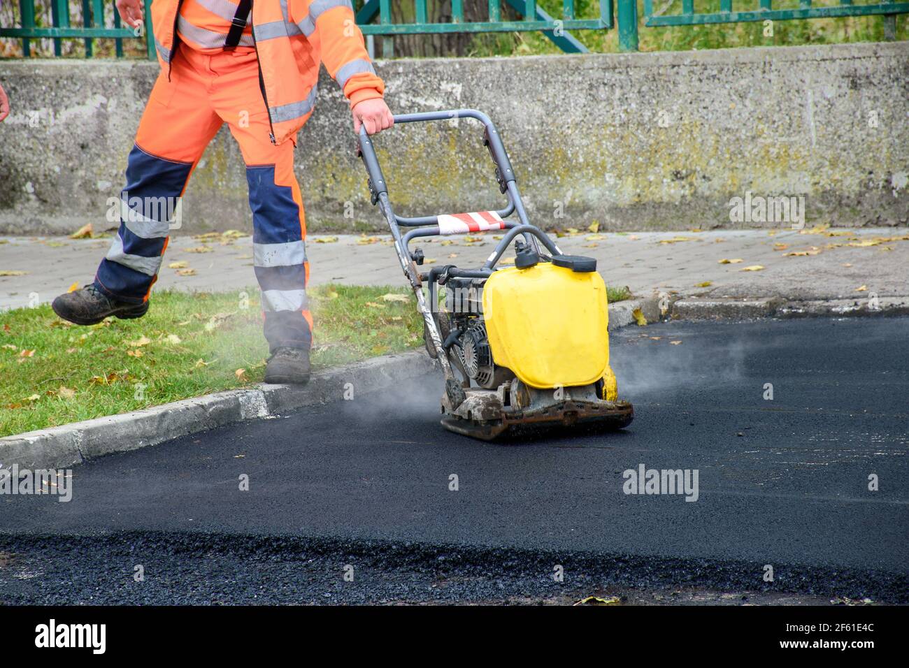 Paving worker uses vibratory plate compactor to compact new asphalt near curbstones Stock Photo