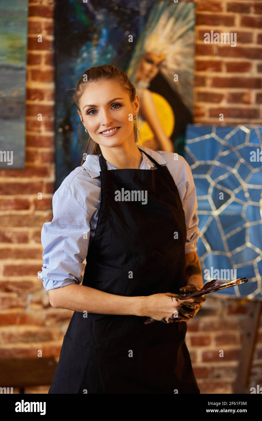 Portrait of young smiling female artist wearing apron in art studio ...
