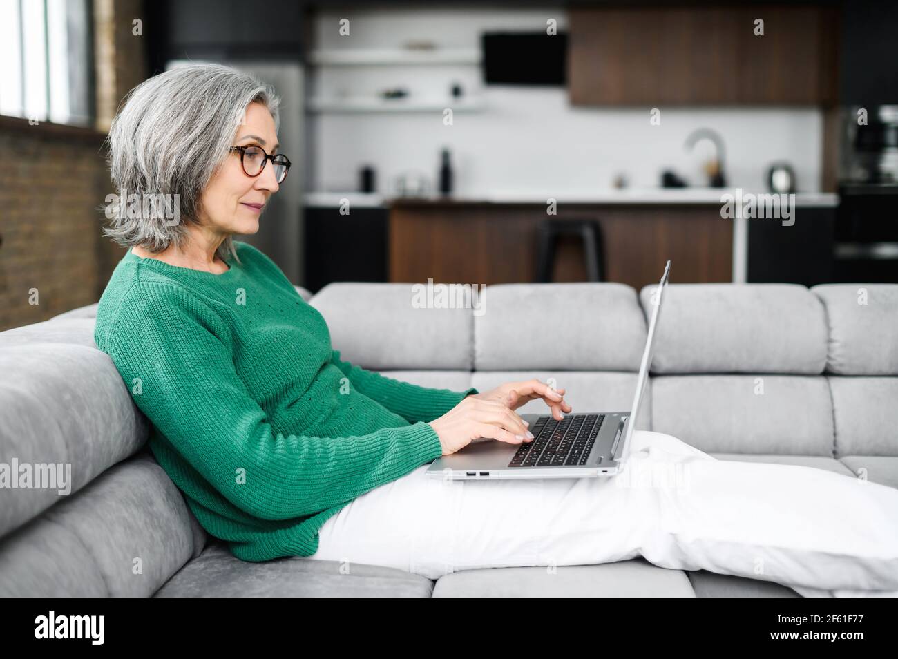 Side view of a senior female with a laptop lying down on the couch at cozy living room. Serious