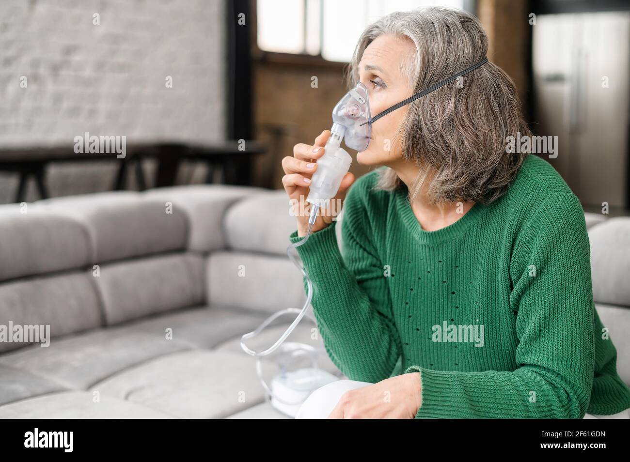 Upset senior woman feeling unwell, has shortness of breath, asthma, using oxygen mask sitting on the couch at home. Sick mature woman makes inhalation with a nebulizer at home, treating herself Stock Photo