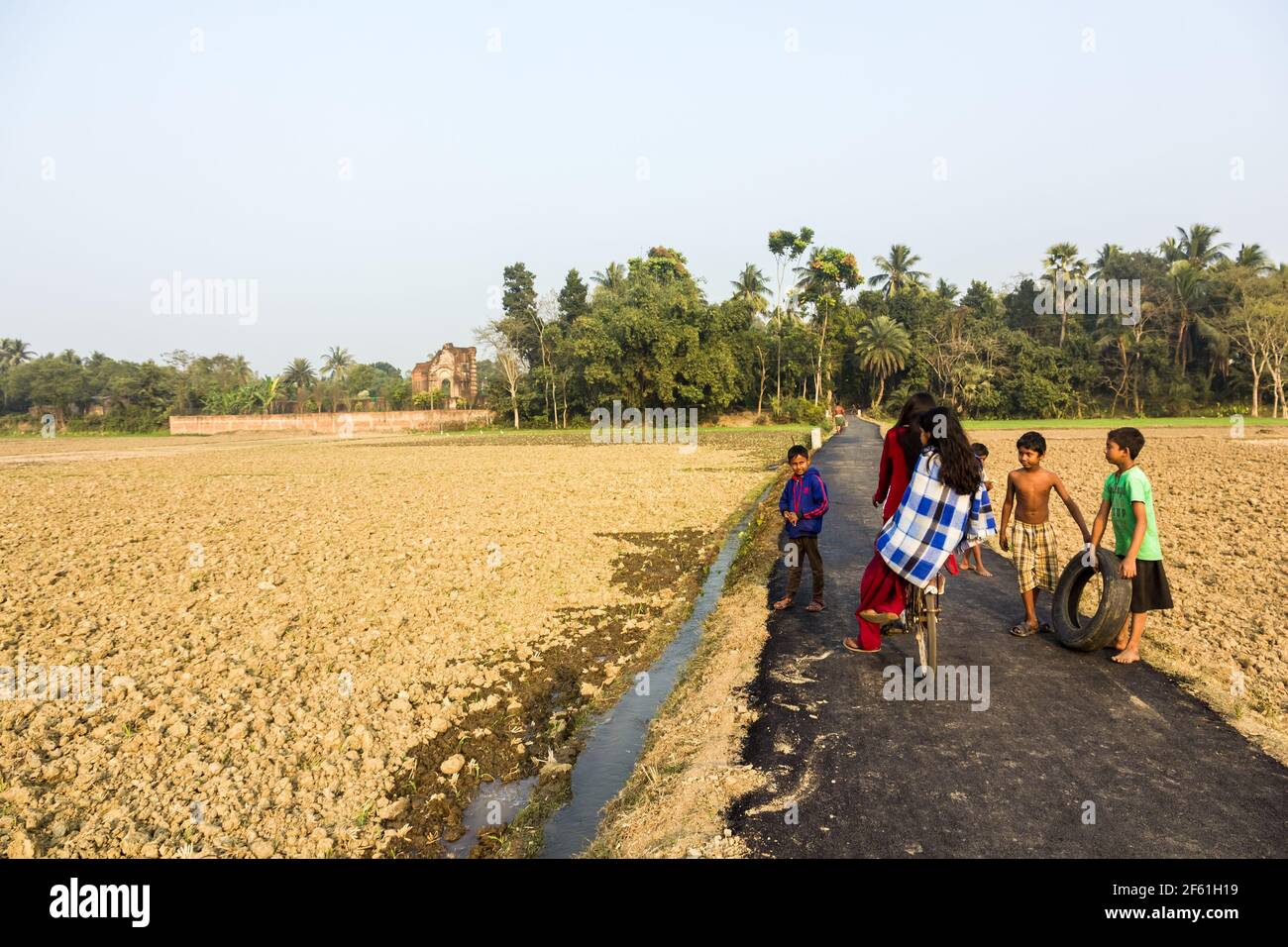 Murshidabad, West Bengal, India - January 2018: Girls riding a bicycle past a group of boys on a rural country road in the fields in the town of Mursh Stock Photo