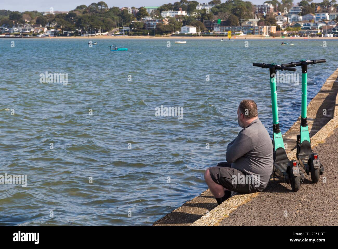 Man sitting on wall with two beryl electric scooters e scooters escooters at Sandbanks, Poole, Dorset UK in March Stock Photo