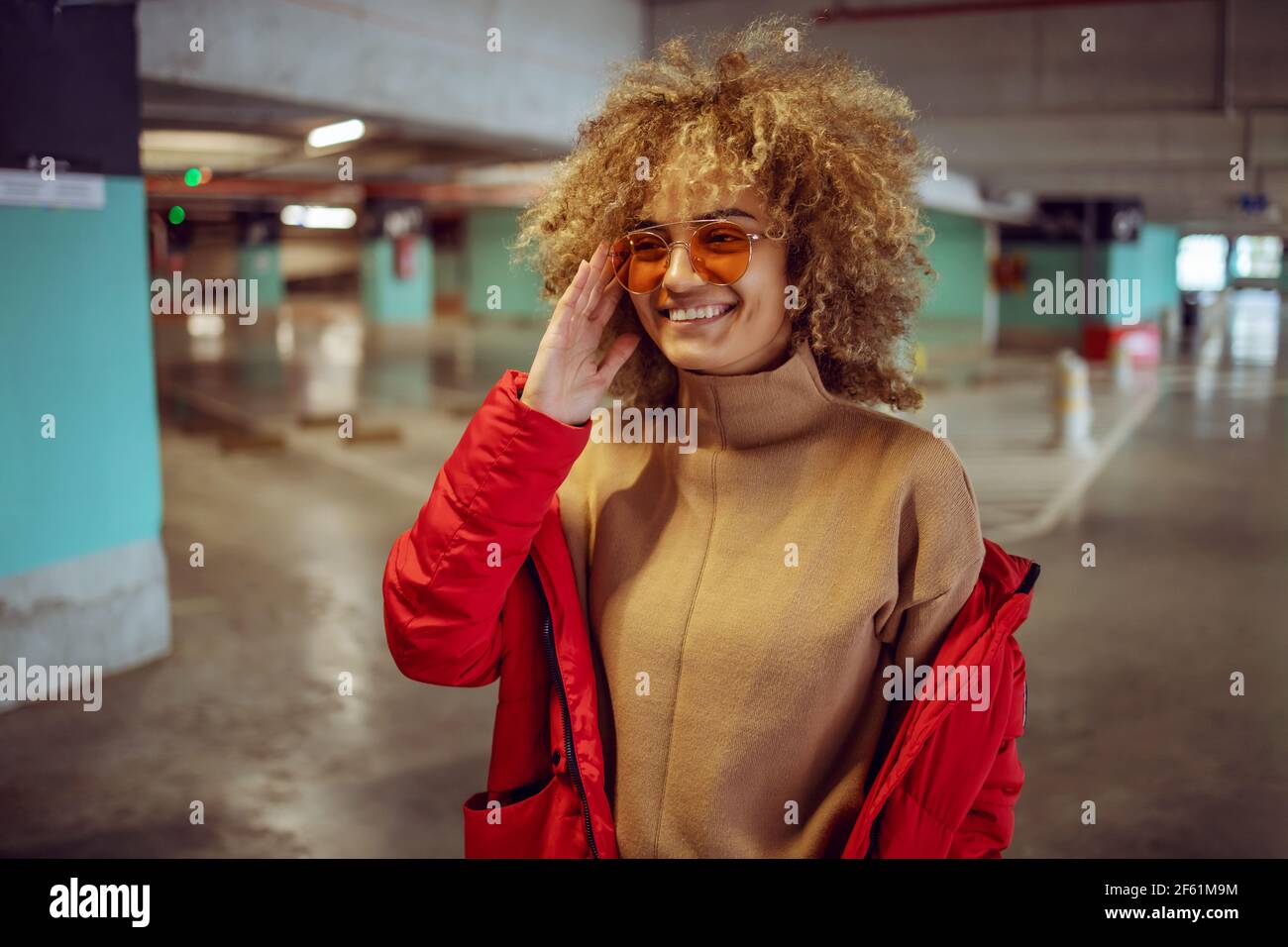 Smiling mixed race hip hop girl in jacket standing in garage and adjusting sunglasses. Stock Photo