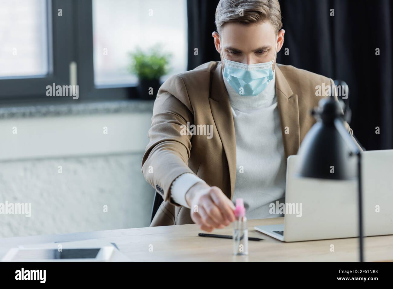 young businessman in medical mask taking antiseptic while sitting at workplace Stock Photo