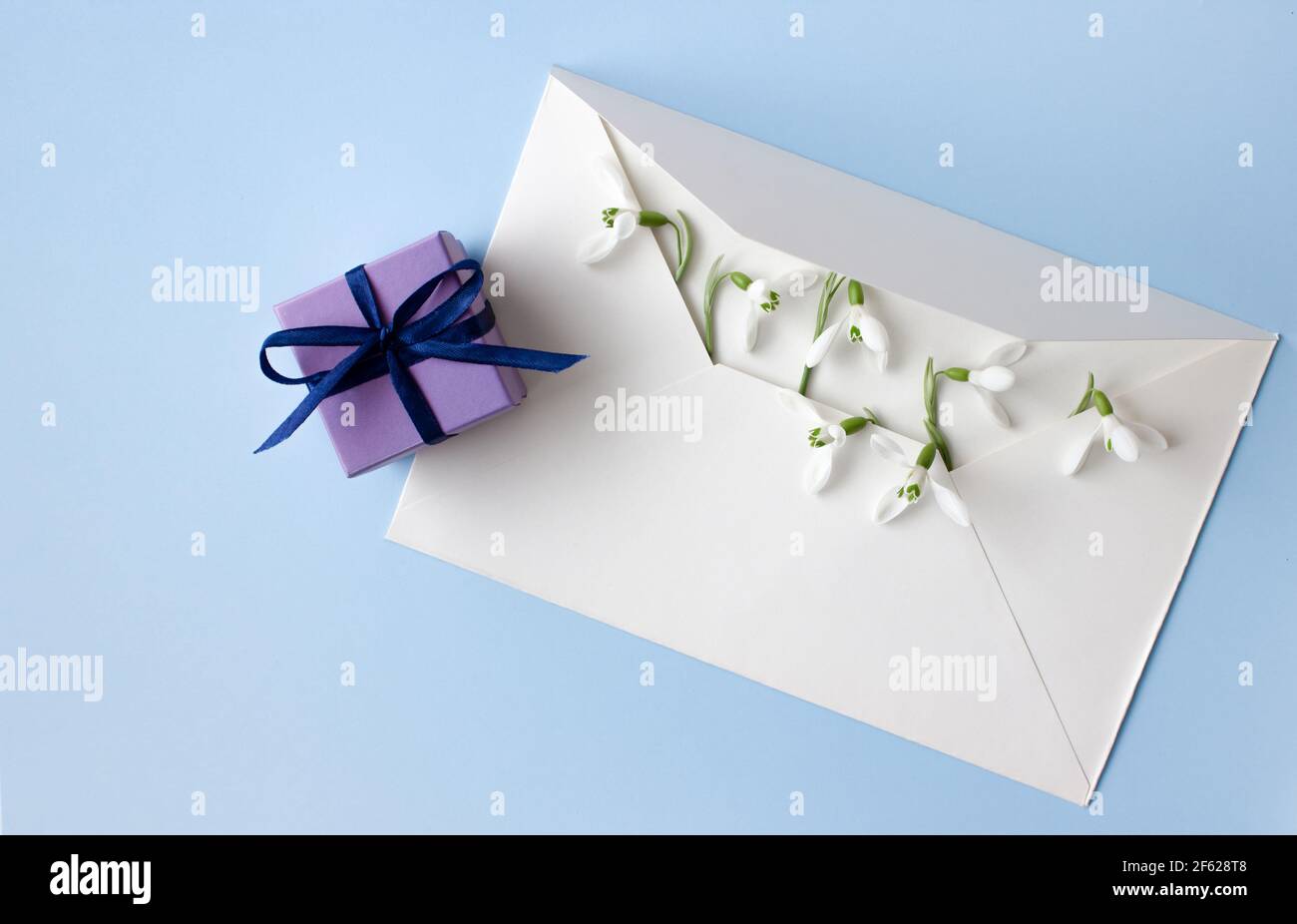 Beautiful snowdrops in white envelope and purple gift box on light blue background, closeup, space for text, top view. Stock Photo